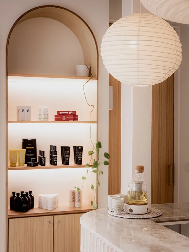 An inviting spa reception area with a curved wooden shelf displaying beauty products under an arched mirror, a paper lantern pendant light, and a tea set on a marble countertop, creating a warm and relaxing atmosphere